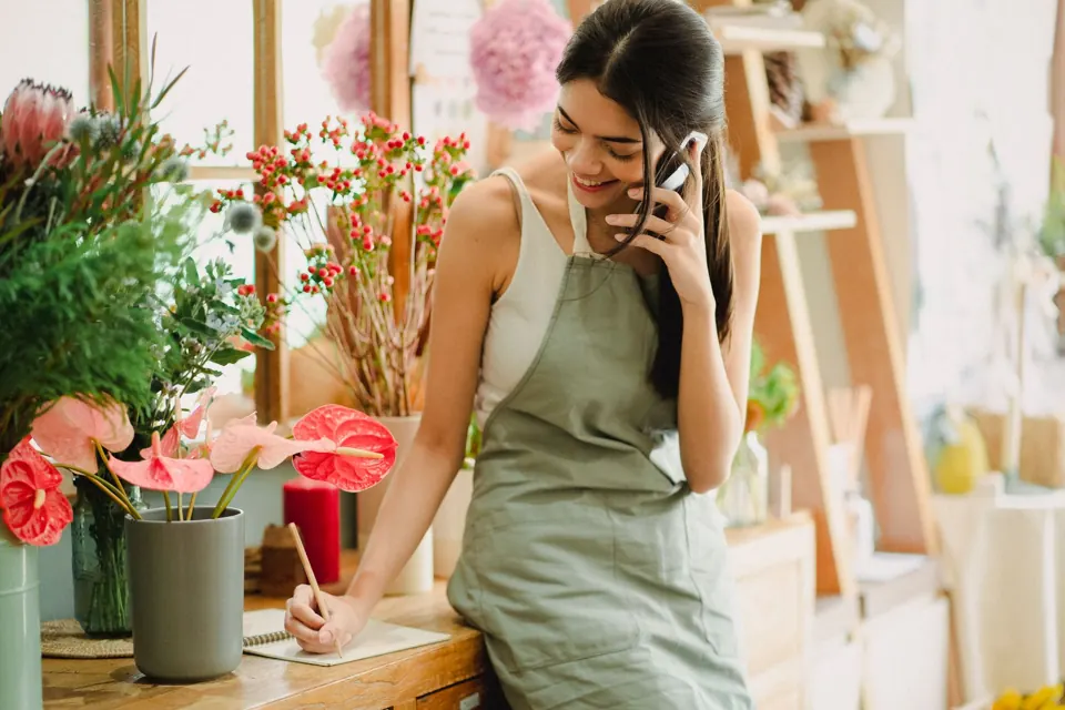 A smiling florist takes a customer call on her phone while writing down an order